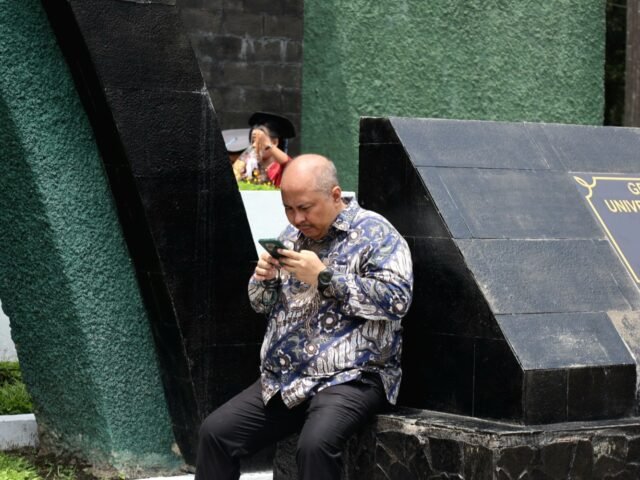 SEJENAK | Seorang pria menjauhi area keramaian sejenak. Ia menyempatkan diri membuka ponsel di tengah hiruk-pikuk acara wisuda, Jumat (29/8/2025). | Imelda Sari Manalu