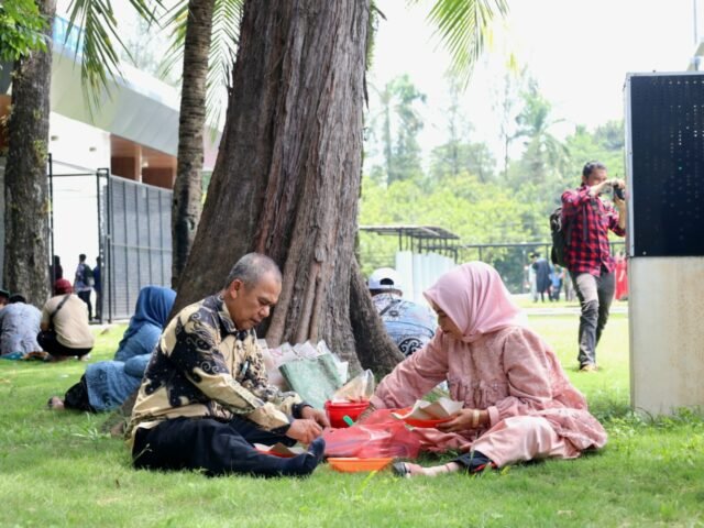 RINDANG | Sambil menunggu sang anak, kedua orang tua memakan bekal yang dibawa dari rumah. Tempatnya sederhana, hanya beralas rumput dan beratap pohon, Jumat (29/8/2025). | Dinar Fazira Fitri