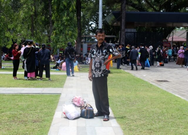 MENJAGA | Seorang ayah berdiri sambil menjaga barang di sekitarnya. Sigap ia lakukan, sambil menunggu wisudawan mengambil foto bersama, Jumat (29/8/2025). | Dinar Fazira Fitri
