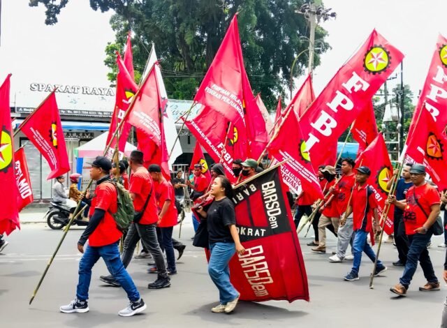 ARAK-ARAK | Massa aksi berjalan beriringan sambil mengibarkan bendera organisasi buruh di sepanjang jalan kota. Deretan bendera merah mencerminkan solidaritas dan semangat perjuangan, Jumat (1/5/2026). | Iyusarah Pakpahan