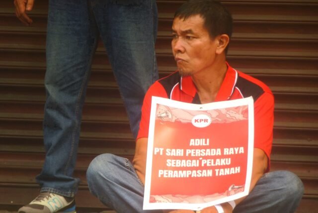 MENUNTUT | Seorang peserta aksi duduk sambil memegang poster berisi tuntutan saat peringatan Hari Buruh. Poster tersebut menyoroti dugaan perampasan tanah, Jumat (1/5/2026). | Sepania Margareta Hutabarat