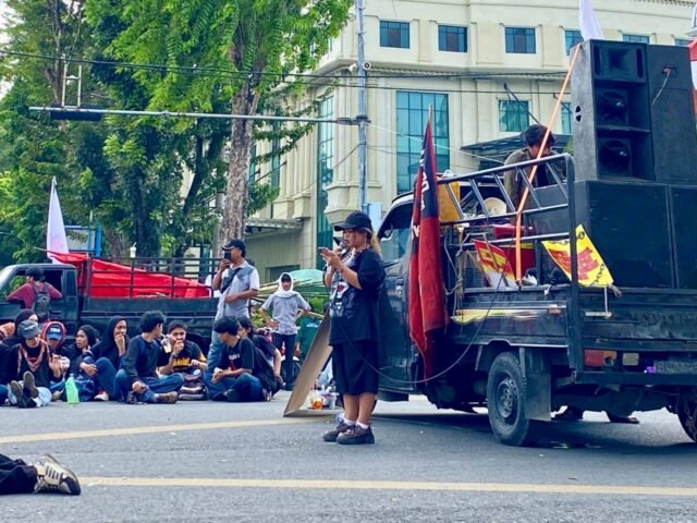 MENYUARAKAN | Seorang peserta aksi menyampaikan orasi di titik aksi. Sejumlah massa duduk di badan jalan sambil mendengarkan, Jumat (1/5/2026). | Dwi Yolanda Naro Situmorang